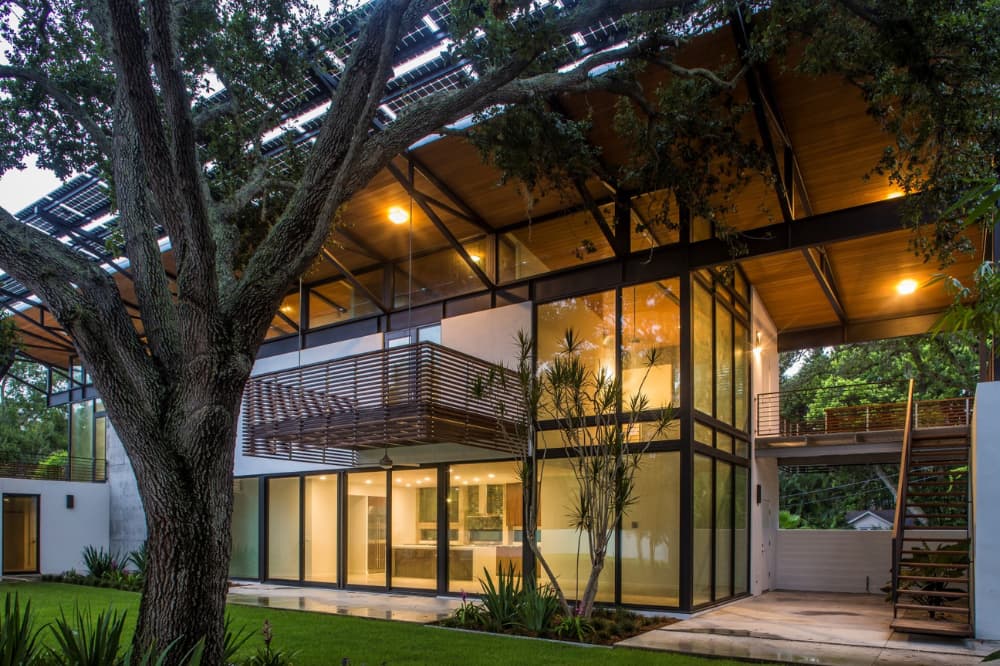 Modern glass-walled house opening onto a lawn beneath deep roof overhangs and mature trees
